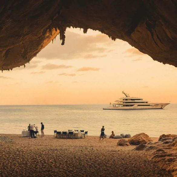 Inside a cave, a yacht is anchored as beachgoers enjoy the sun on the shore.